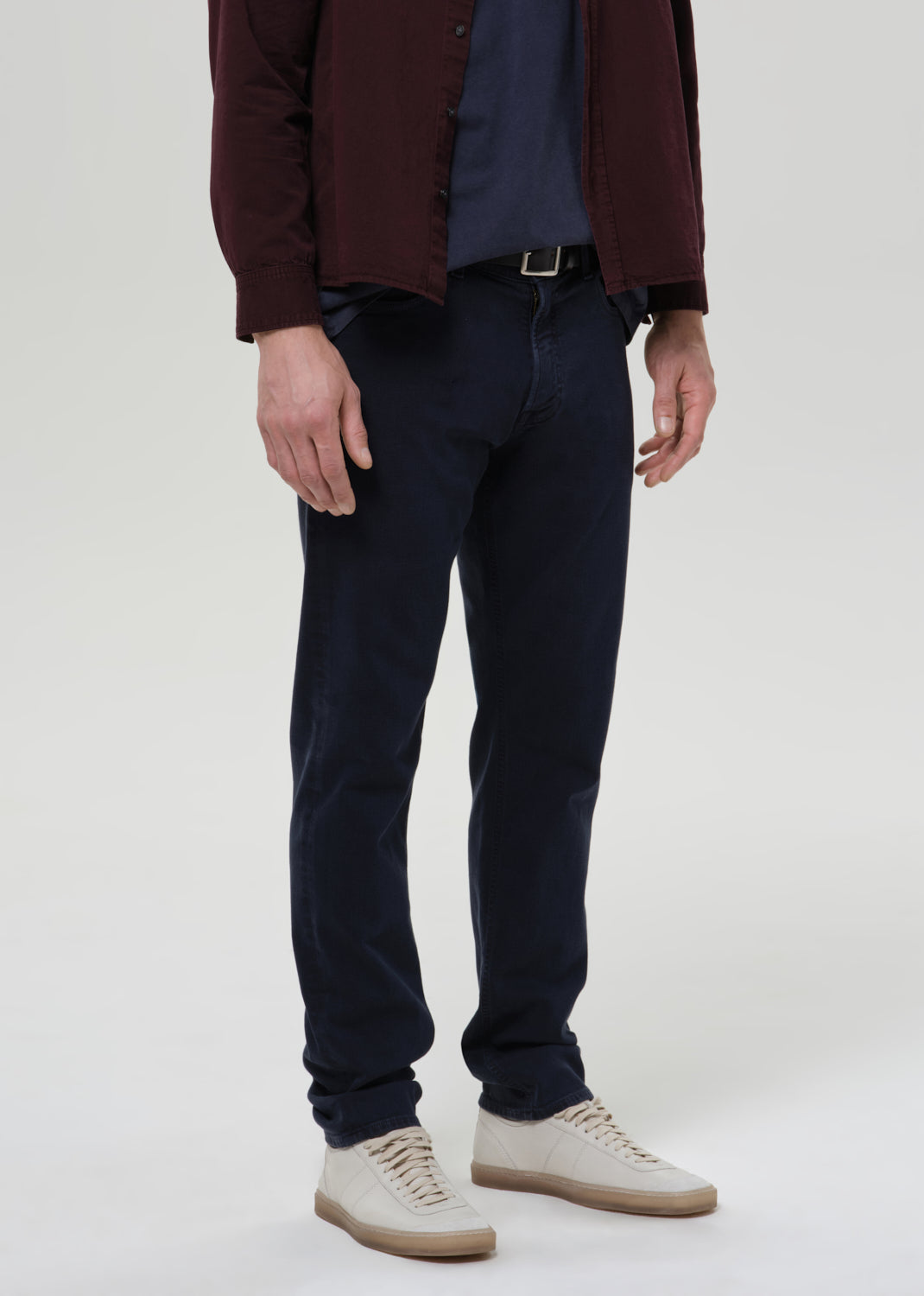 A man wearing dark navy slim-fit jeans, a dark blue t-shirt, a maroon button-up shirt, and beige sneakers, shown from the waist down against a grey background.