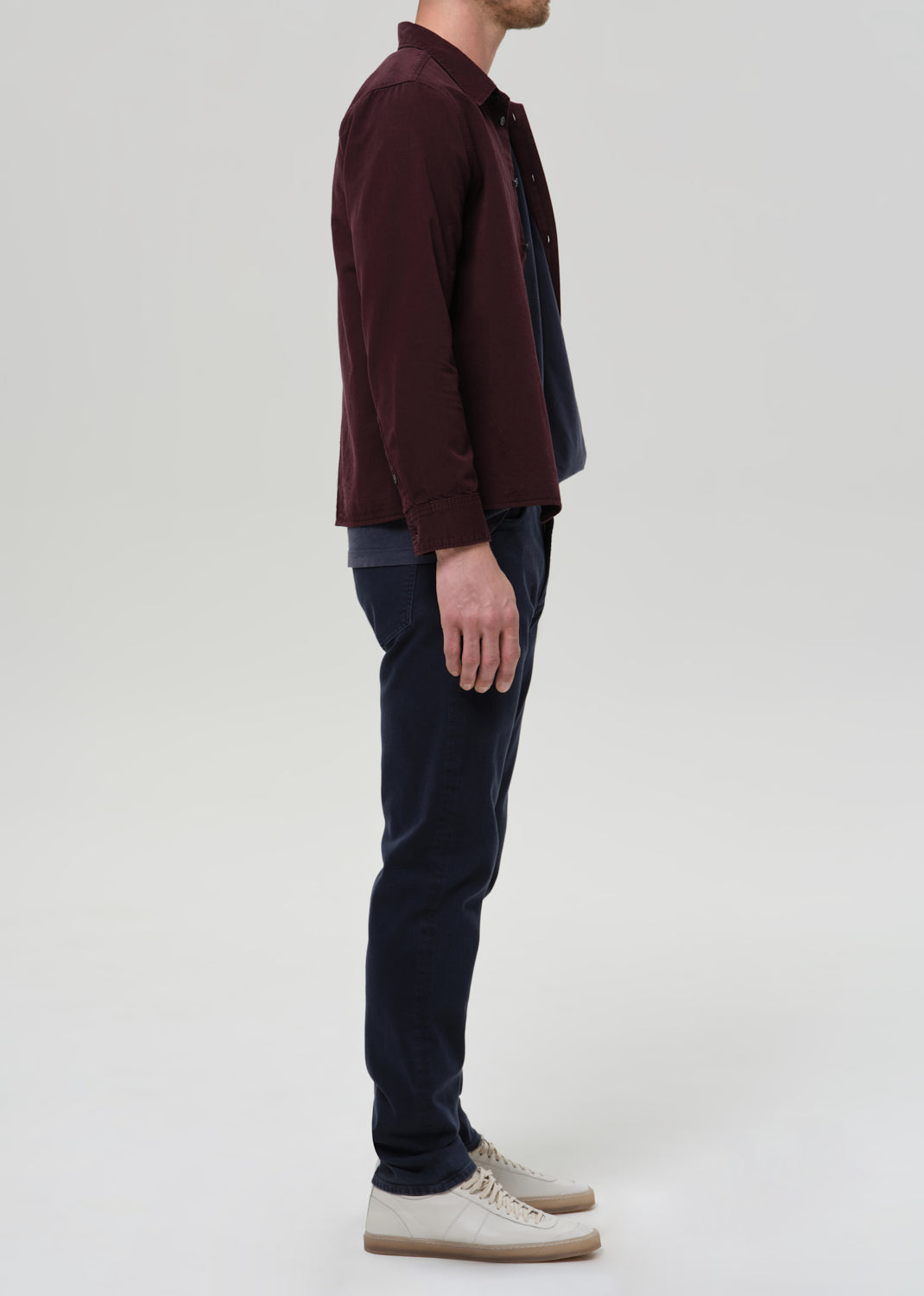 A man shown in profile wearing a dark burgundy overshirt over a navy shirt, navy trousers, and beige sneakers, photographed against a grey background.