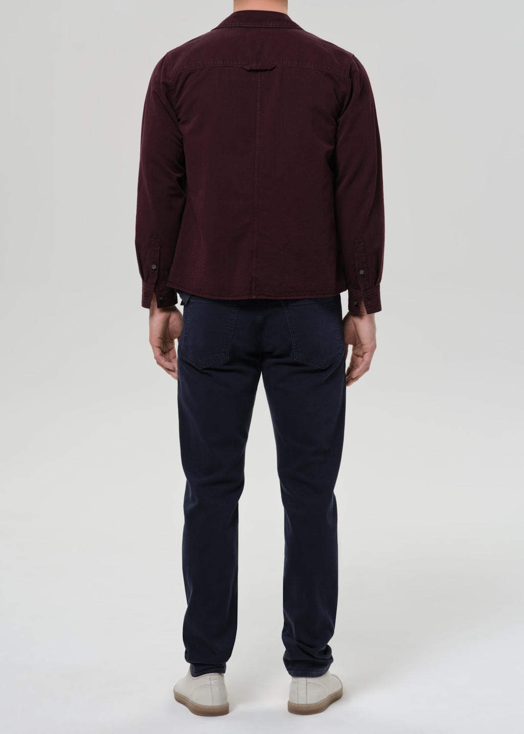 A man viewed from the back wearing a dark burgundy long-sleeve button-up shirt, navy blue pants, and off-white shoes, standing against a grey background.