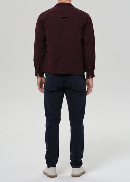 A man viewed from the back wearing a dark burgundy long-sleeve button-up shirt, navy blue pants, and off-white shoes, standing against a grey background.