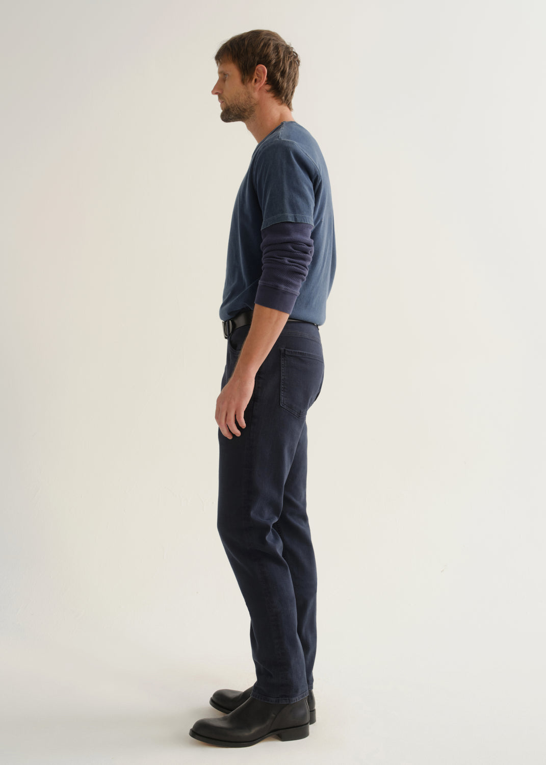 Person wearing dark navy pants, a blue long sleeve shirt, and black boots, side full body view on a light grey background.