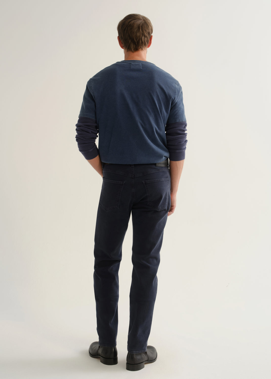 Person wearing dark navy pants, a blue long sleeve shirt, and black boots, back full body view on a light grey background.