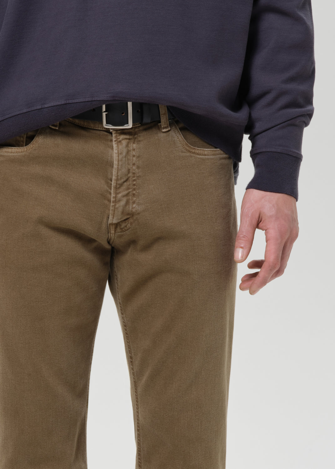 A close-up of a person wearing brown chino pants with a black belt and a dark navy sweatshirt, shown from the chest down against a grey background.