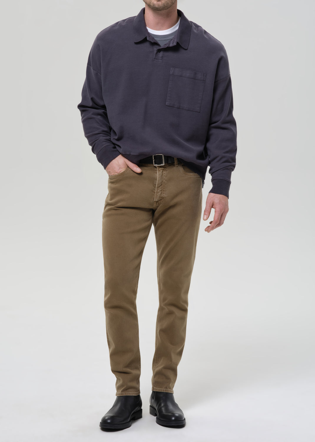 A man wearing a dark navy long-sleeve polo shirt, olive brown trousers, and black leather boots, standing against a grey background.