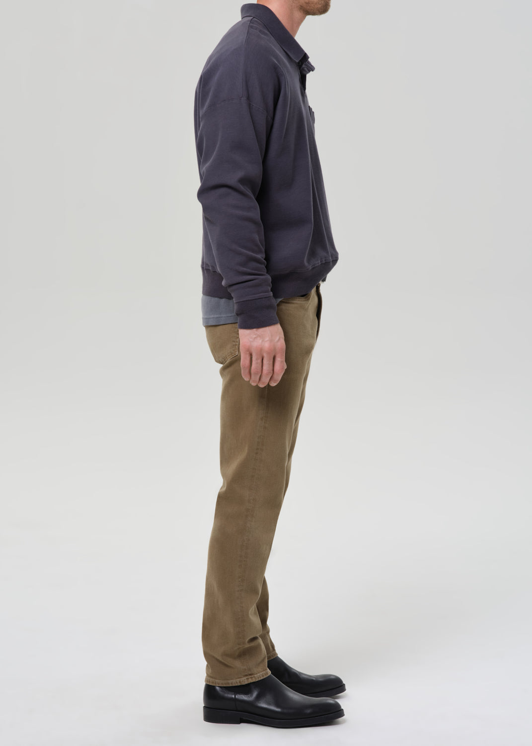 A man shown in profile wearing a dark navy long-sleeve button-up shirt, olive-brown trousers, and black leather shoes, standing against a grey background.