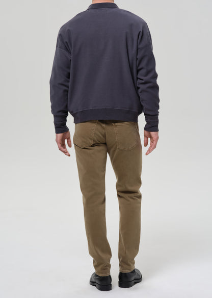 A man viewed from behind wearing a dark navy crewneck sweatshirt, olive brown pants, and black shoes, standing against a grey background.
