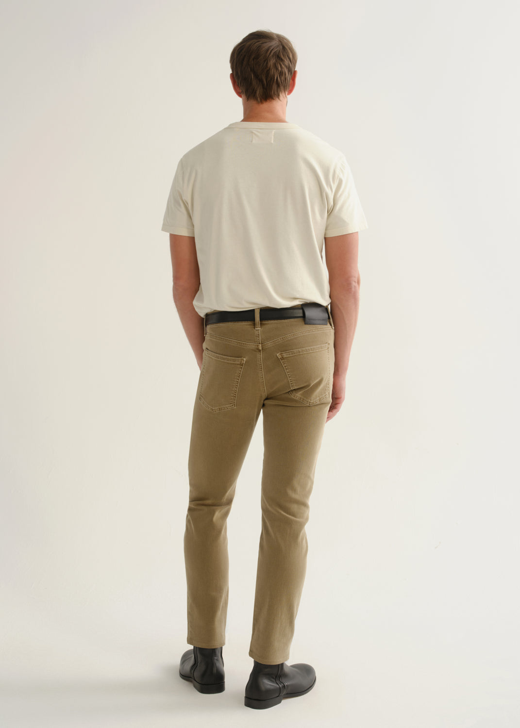 Person wearing khaki brown pants, a white short sleeve t-shirt, and black boots, back full body view on a light grey background.