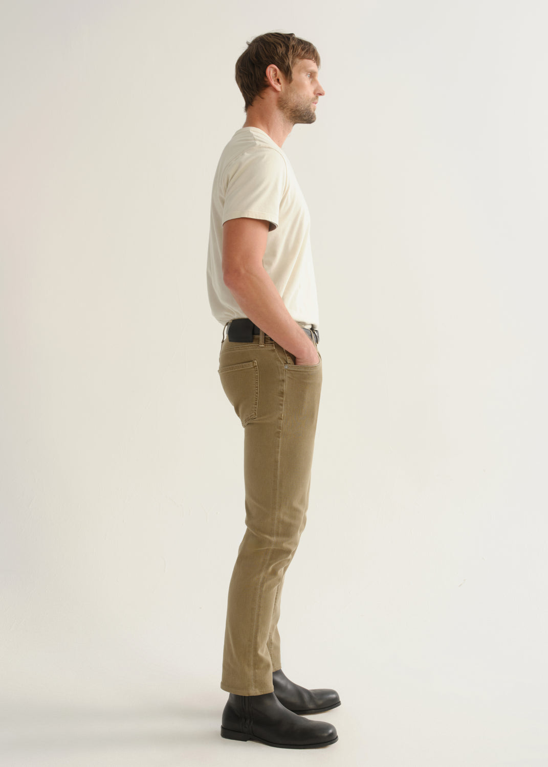 Person wearing khaki brown pants, a white short sleeve t-shirt, and black boots, side full body view on a light grey background.