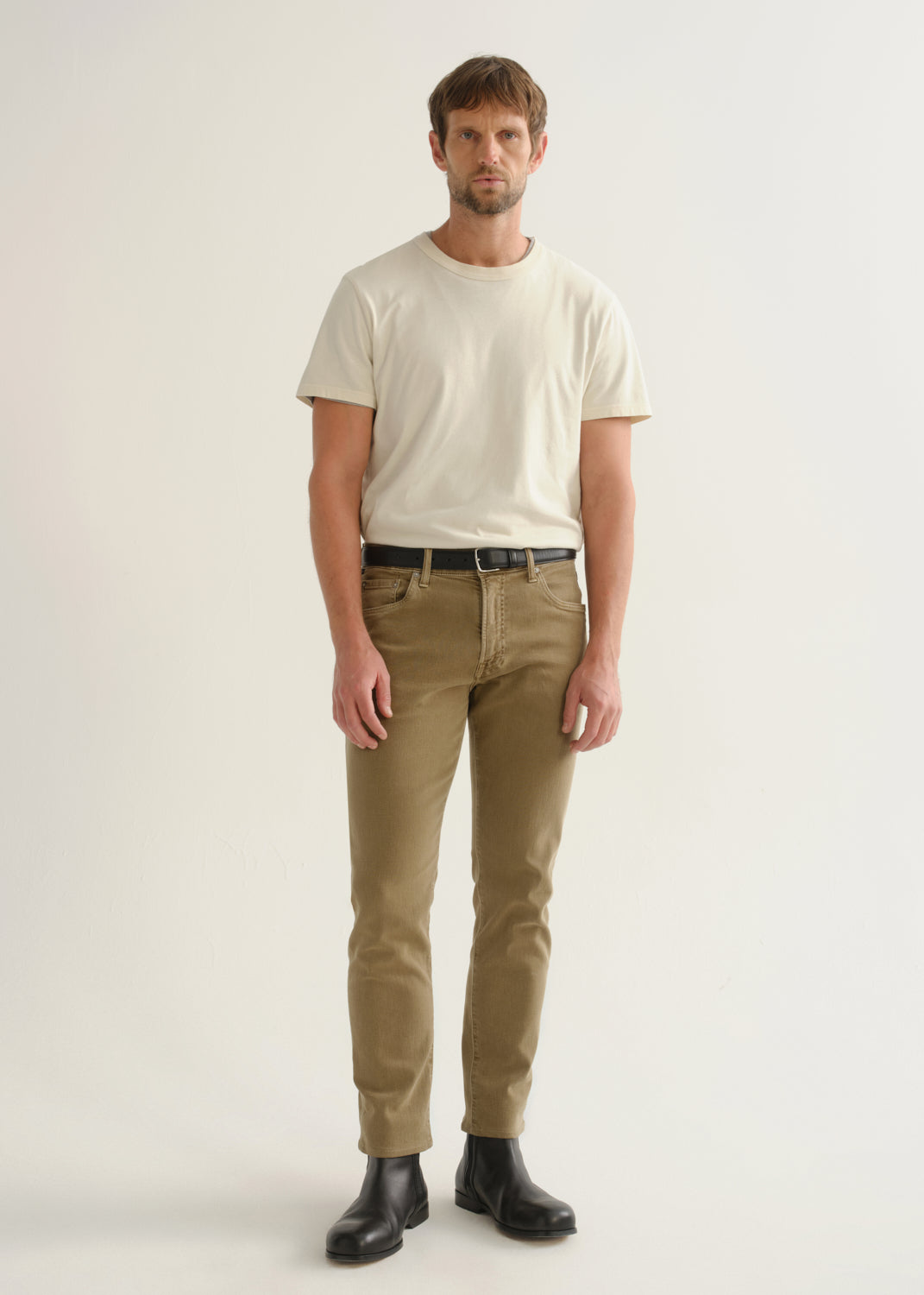 Person wearing khaki brown pants, a white short sleeve t-shirt, and black boots, front full body view on a light grey background.