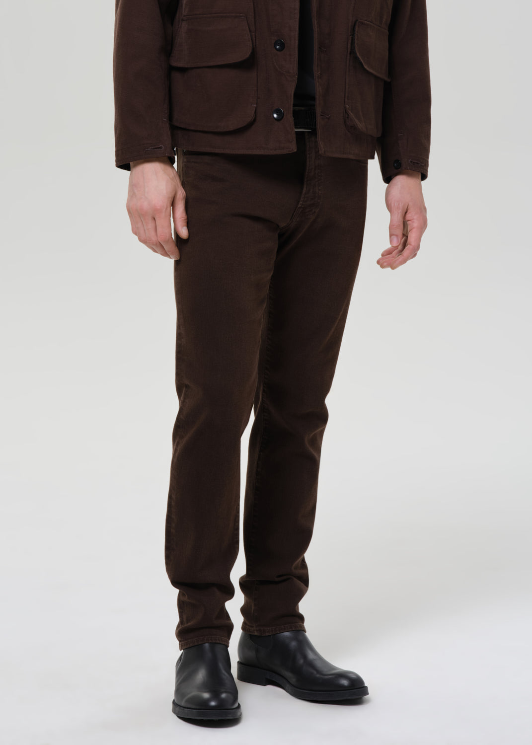 Person wearing a brown jacket and pants with black shoes on a grey background