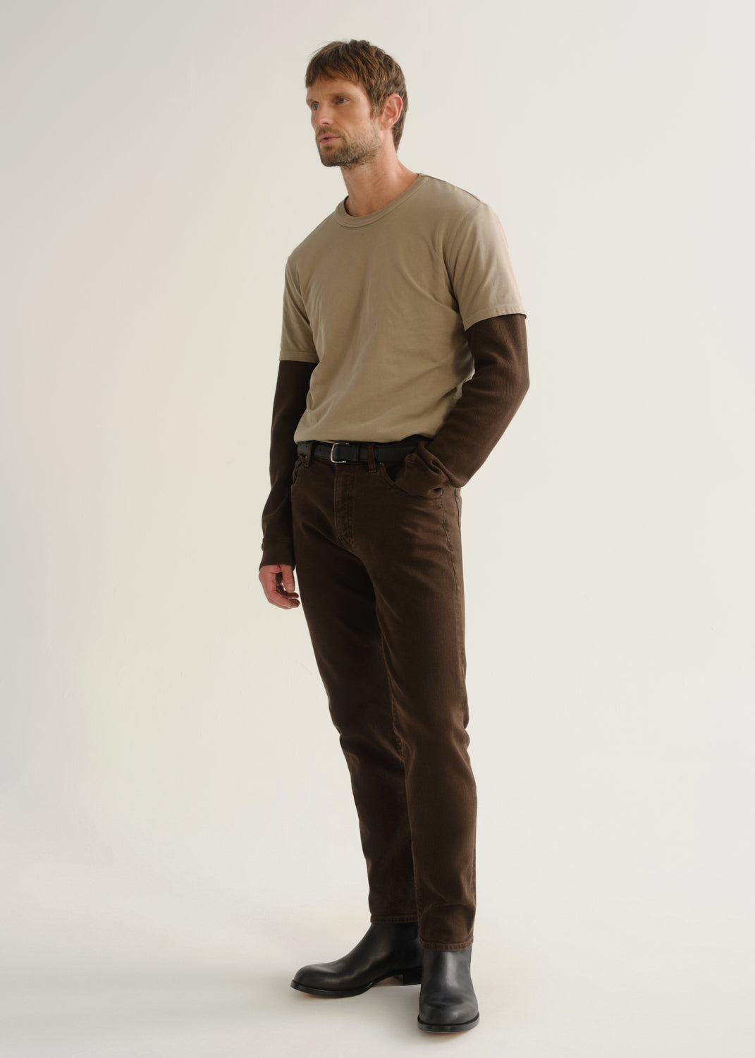 Person wearing dark brown jeans, a beige short sleeve t-shirt layered over a dark brown long sleeve shirt, and black boots, angled front full body view on a light grey background.