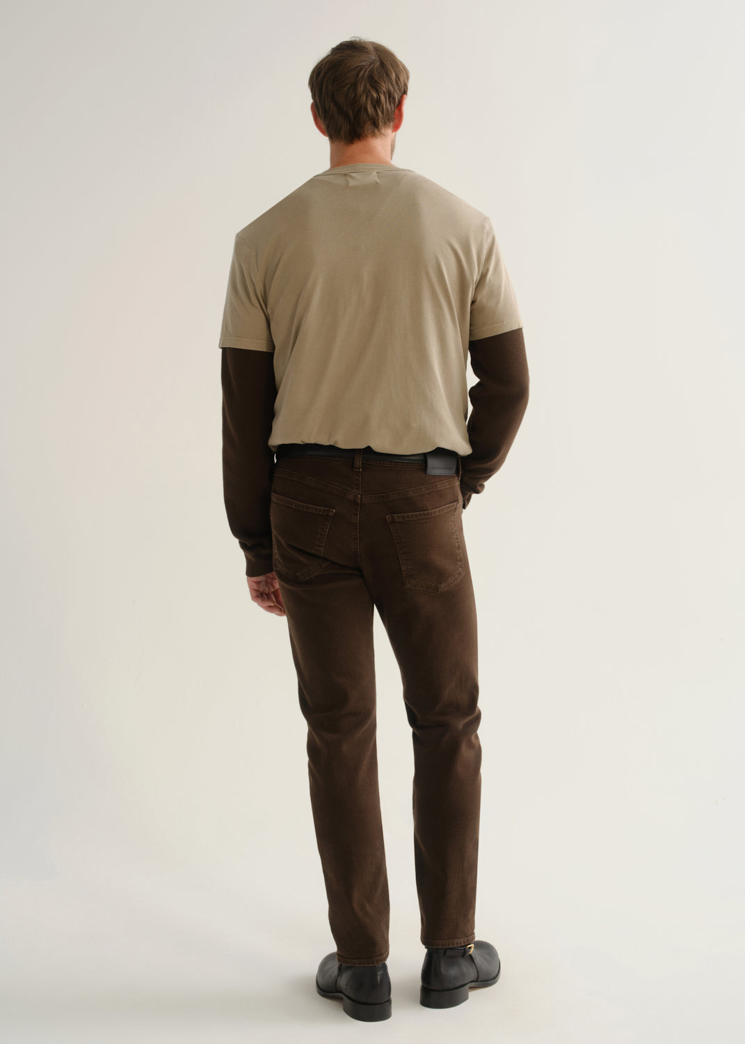 Person wearing dark brown jeans, a beige short sleeve t-shirt layered over a dark brown long sleeve shirt, and black boots, back full body view on a light grey background.