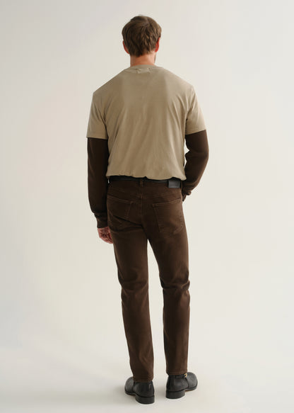 Person wearing dark brown jeans, a beige short sleeve t-shirt layered over a dark brown long sleeve shirt, and black boots, back full body view on a light grey background.