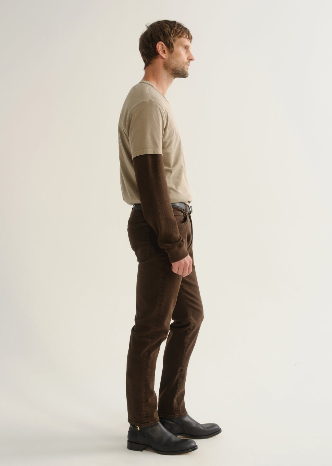 Person wearing dark brown jeans, a beige short sleeve t-shirt layered over a dark brown long sleeve shirt, and black boots, side full body view on a light grey background.