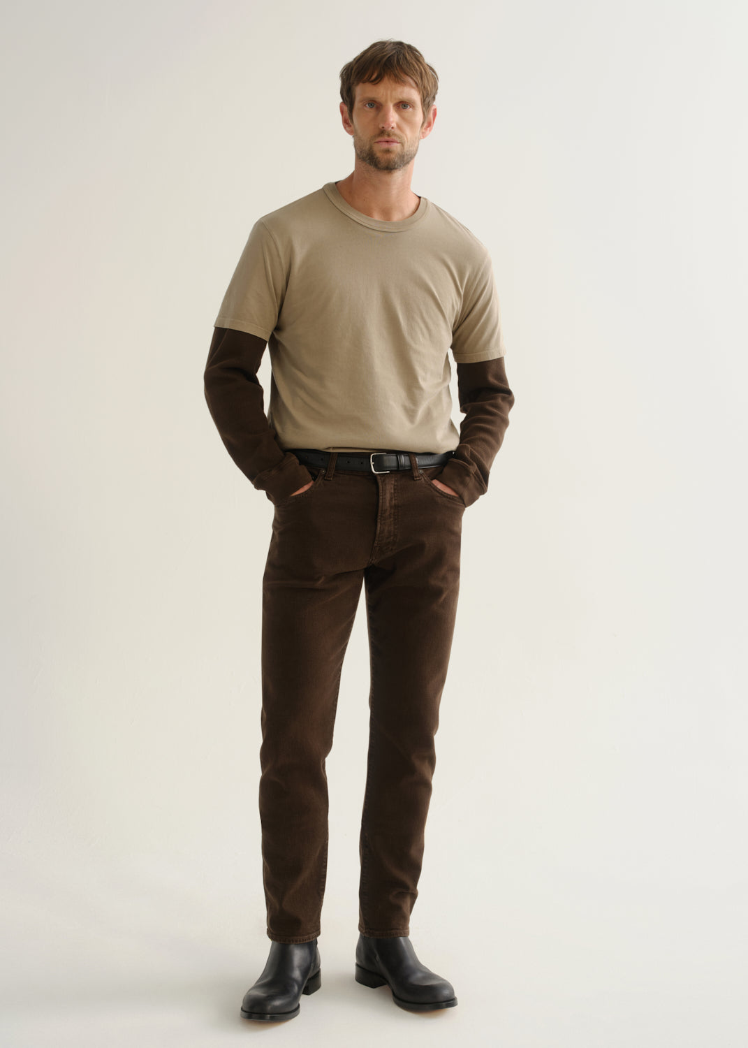 Person wearing dark brown jeans, a beige short sleeve t-shirt layered over a dark brown long sleeve shirt, and black boots, front full body view on a light grey background.