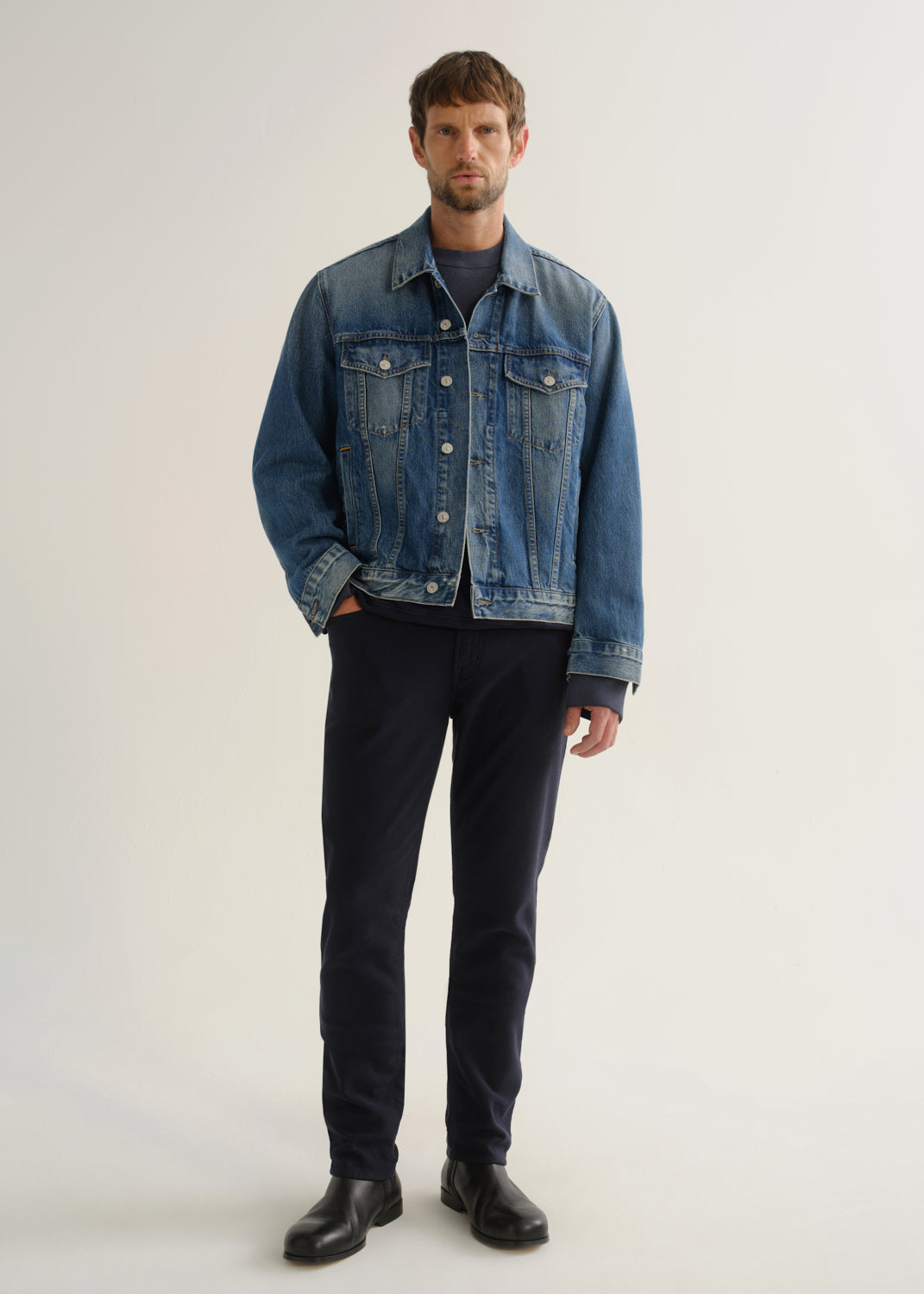 Person wearing dark indigo blue jeans, a medium blue denim jacket, a black shirt, and black boots, front full body view on a light grey background.