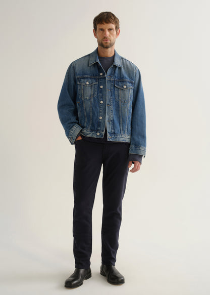 Person wearing dark indigo blue jeans, a medium blue denim jacket, a black shirt, and black boots, front full body view on a light grey background.