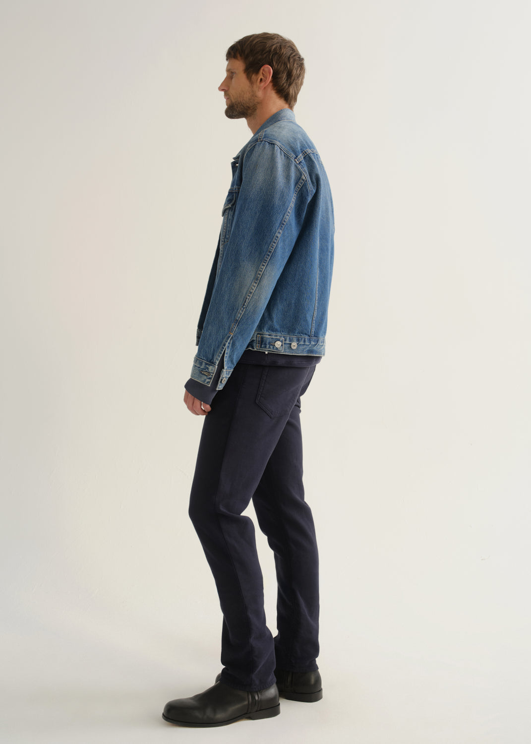 Person wearing dark indigo blue jeans, a medium blue denim jacket, and black boots, side full body view on a light grey background.