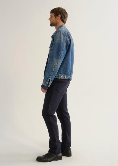 Person wearing dark indigo blue jeans, a medium blue denim jacket, and black boots, side full body view on a light grey background.