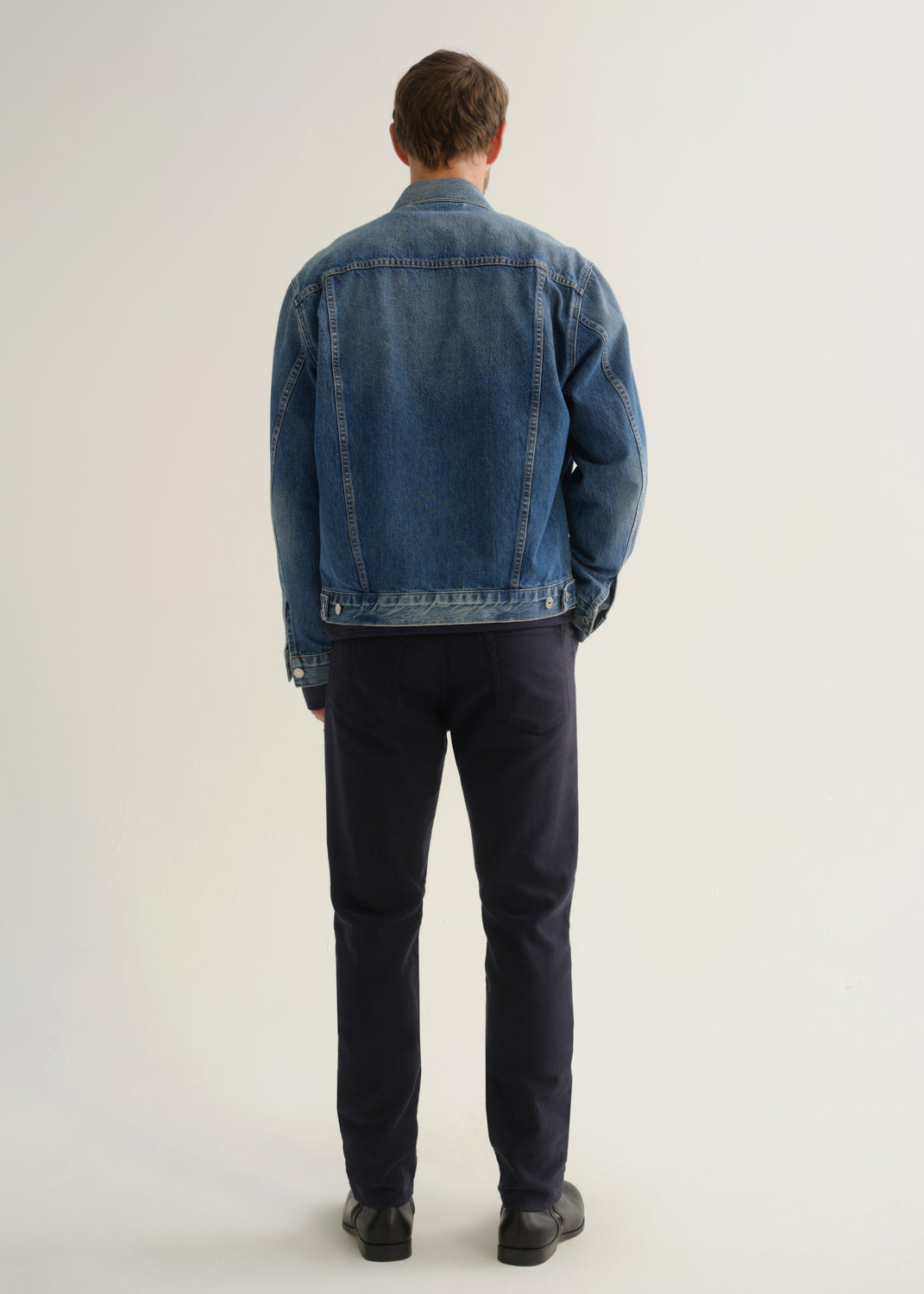 Person wearing dark indigo blue jeans, a medium blue denim jacket, and black boots, back full body view on a light grey background.