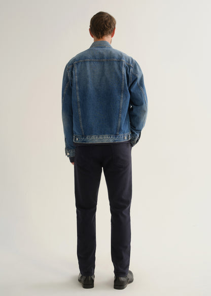 Person wearing dark indigo blue jeans, a medium blue denim jacket, and black boots, back full body view on a light grey background.