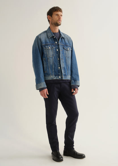 Person wearing dark indigo blue jeans, a medium blue denim jacket, and black boots, angled front full body view on a light grey background.