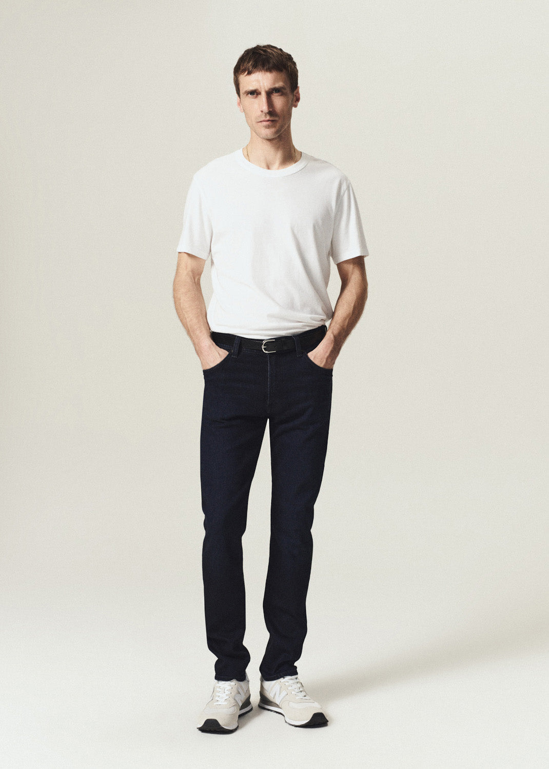 A man modeling a white crew-neck t-shirt, dark navy slim-fit jeans, a black belt, and white sneakers, standing against a light beige studio background.