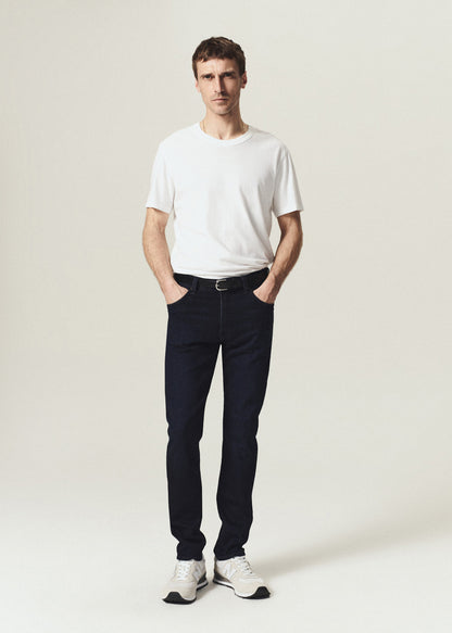 A man modeling a white crew-neck t-shirt, dark navy slim-fit jeans, a black belt, and white sneakers, standing against a light beige studio background.