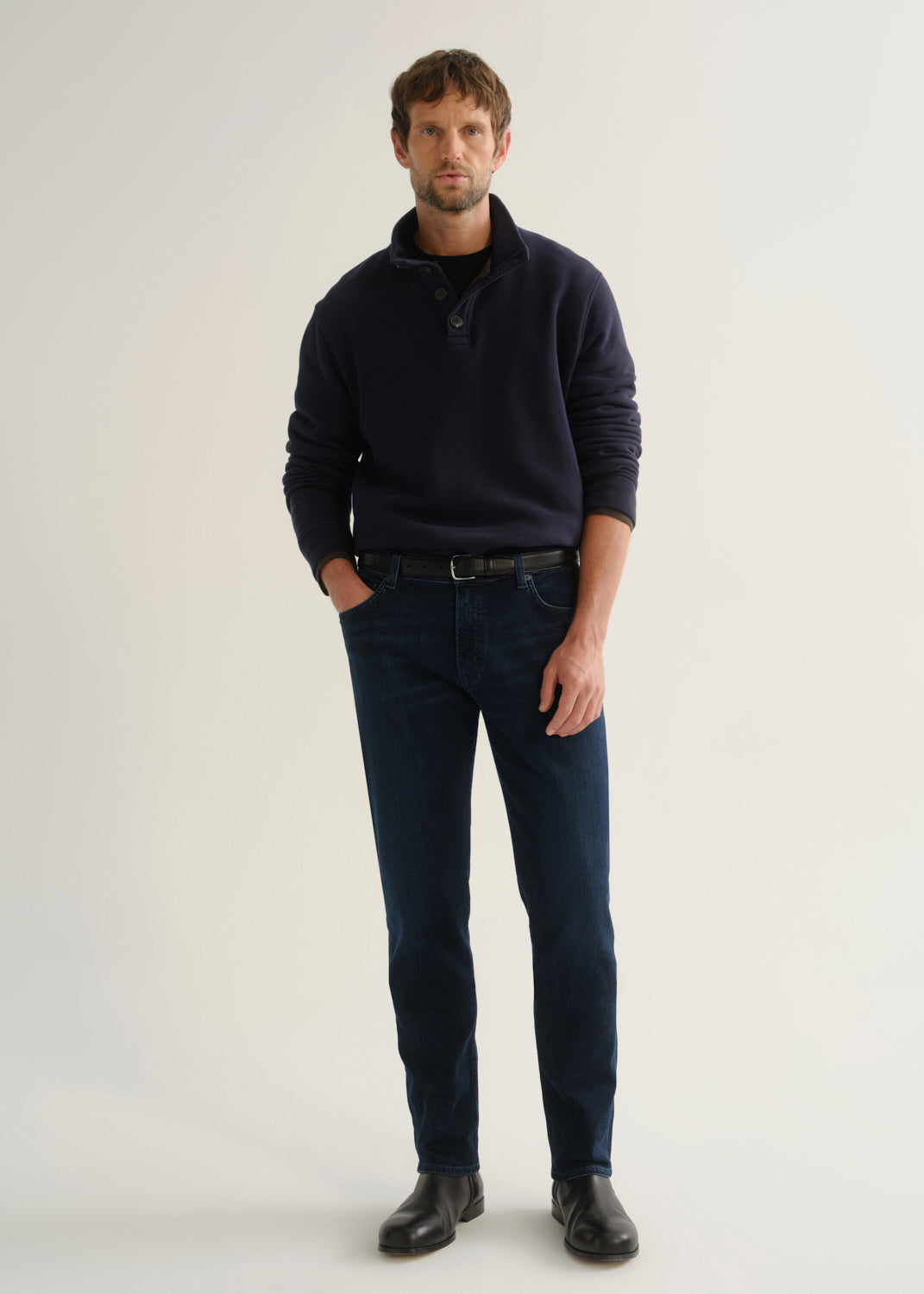 Person wearing dark indigo blue jeans, a navy long sleeve shirt, and black boots, front full body view on a light grey background.