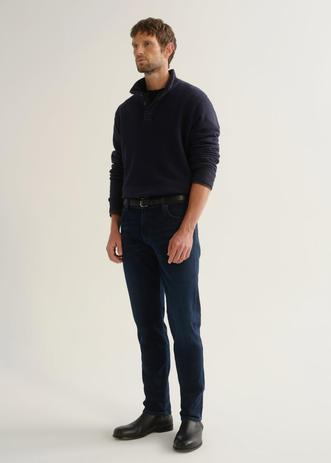 Person wearing dark indigo blue jeans, a navy long sleeve shirt, and black boots, angled front full body view on a light grey background.