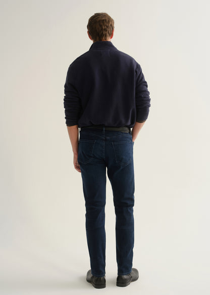 Person wearing dark indigo blue jeans, a navy long sleeve shirt, and black boots, back full body view on a light grey background.
