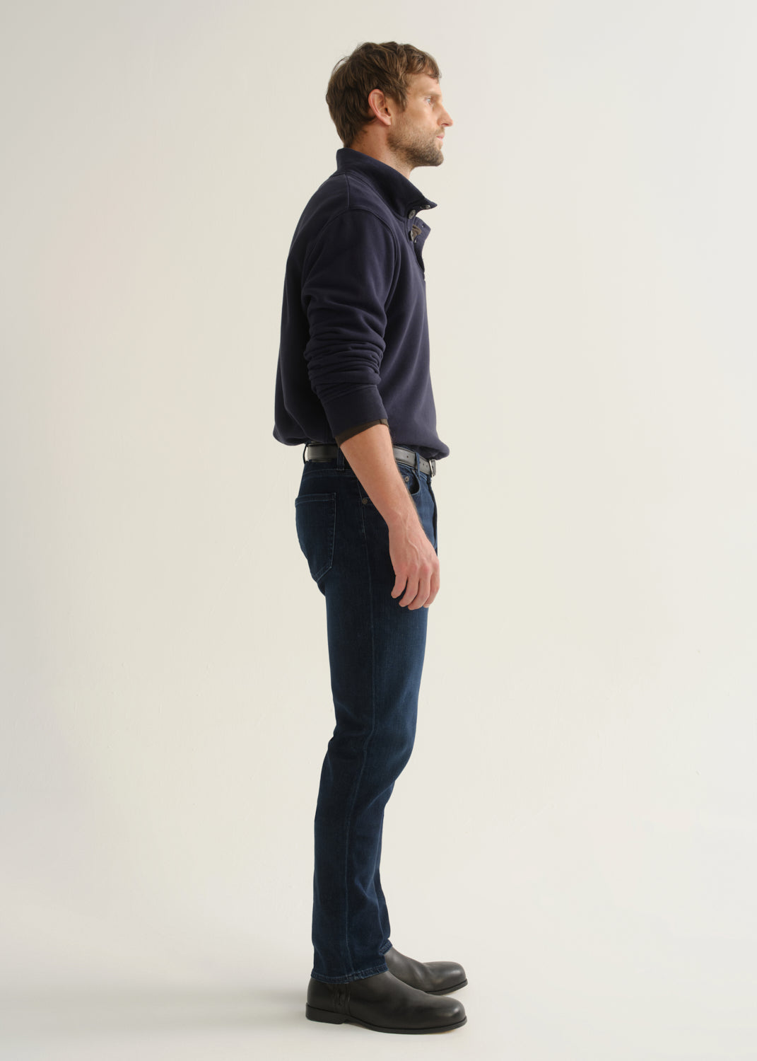 Person wearing dark indigo blue jeans, a navy long sleeve shirt, and black boots, side full body view on a light grey background.