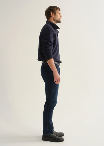 Person wearing dark indigo blue jeans, a navy long sleeve shirt, and black boots, side full body view on a light grey background.