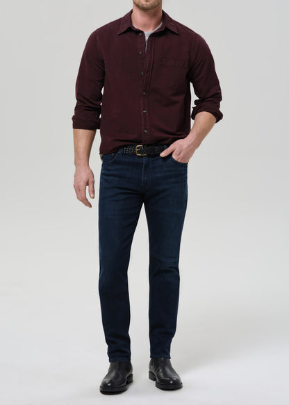 Person wearing dark indigo straight leg jeans with a burgundy button-up shirt on a grey background.