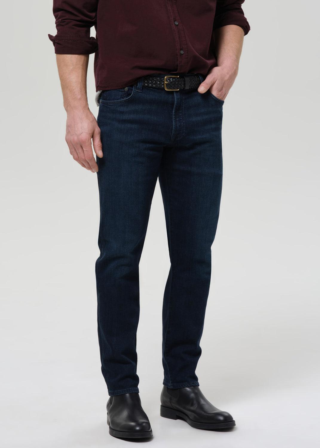 Person wearing dark indigo straight leg jeans with a burgundy button-up shirt on a grey background.