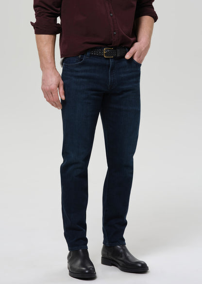 Person wearing dark indigo straight leg jeans with a burgundy button-up shirt on a grey background.