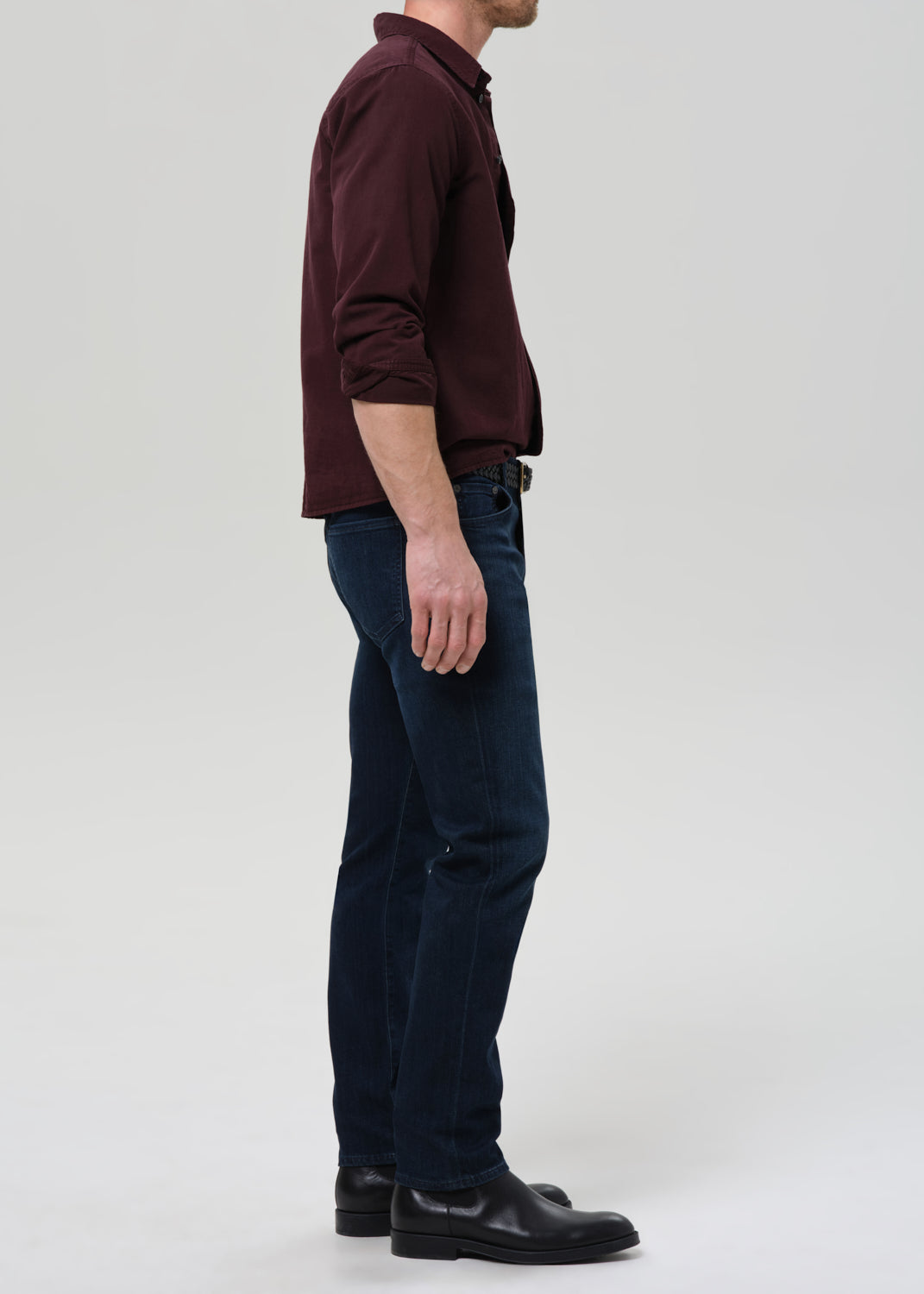 Person wearing dark indigo straight leg jeans with a burgundy button-up shirt viewed from the side on a grey background.