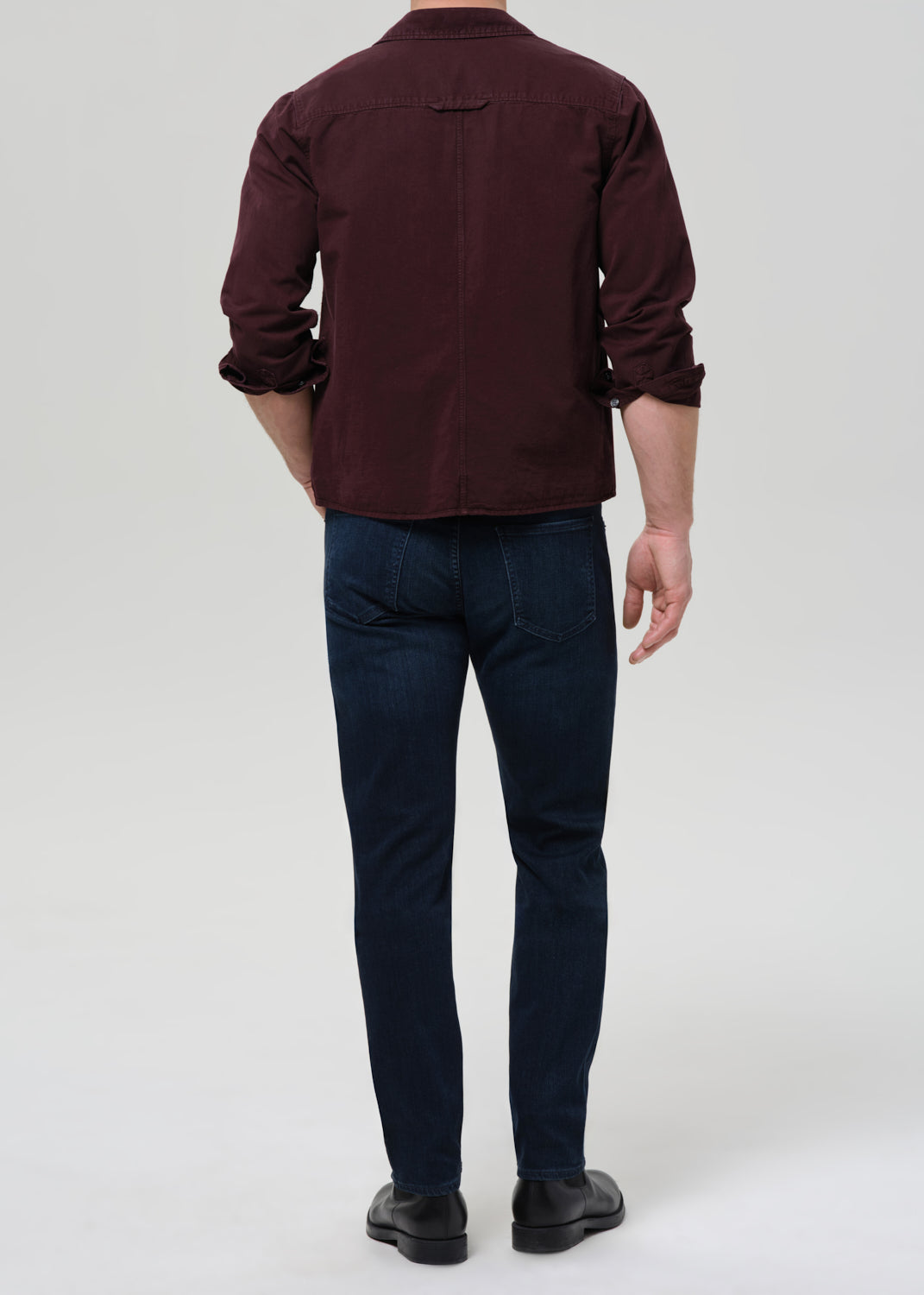 Person wearing dark indigo straight leg jeans with a burgundy button-up shirt viewed from the back on a grey background.