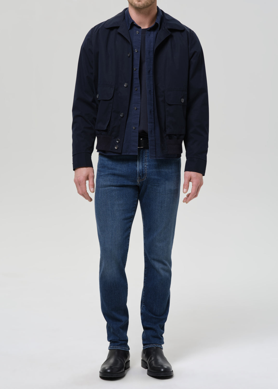 A man wearing a dark navy blue utility jacket over a matching navy shirt, blue jeans, and black leather boots, standing against a grey background.