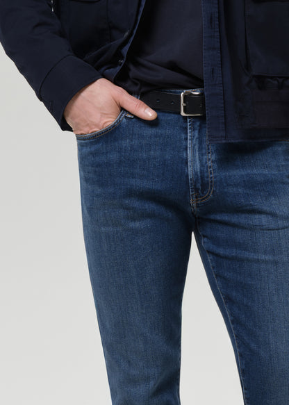 Close-up of a person wearing dark blue denim jeans with a black belt and dark navy jacket, shown from the chest down against a grey background.