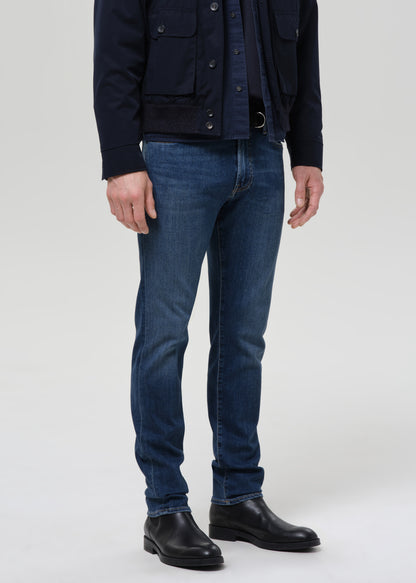 A model from the chest down wearing dark blue slim-fit jeans, a dark navy jacket, a navy shirt, and black leather boots, standing against a grey background.