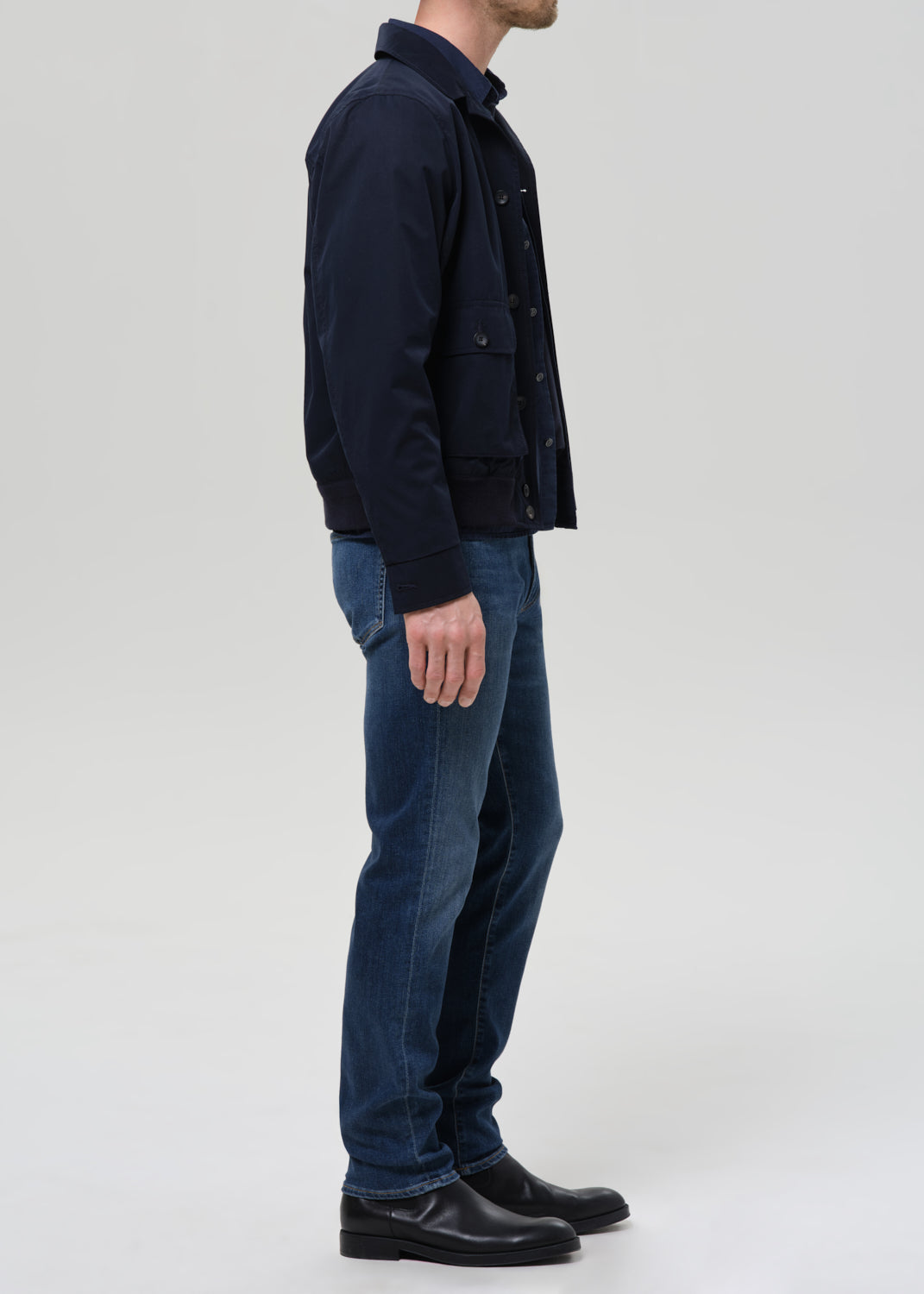 A man shown in profile wearing a dark navy jacket, blue jeans, and black shoes against a grey background.