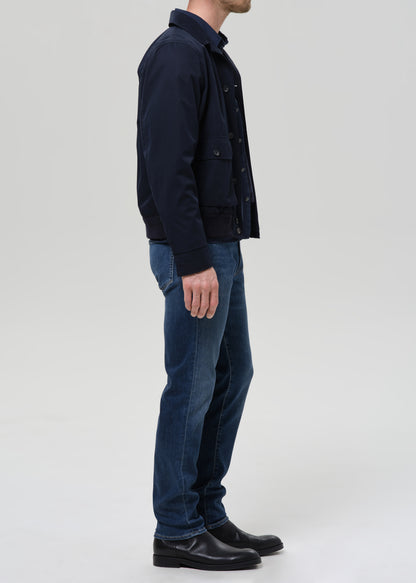 A man shown in profile wearing a dark navy jacket, blue jeans, and black shoes against a grey background.