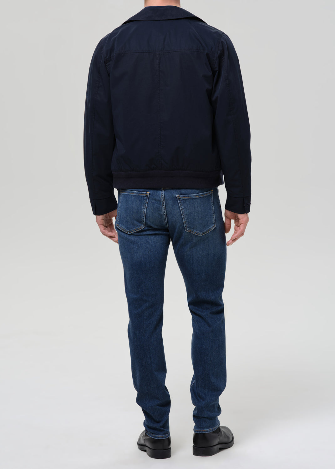 A man viewed from the back wearing a dark navy jacket and blue jeans, standing against a grey background.