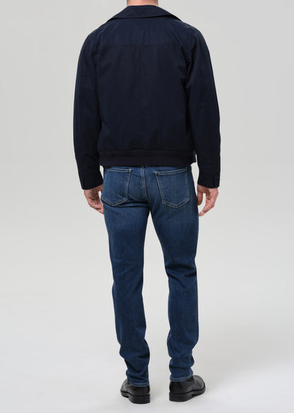 A man viewed from the back wearing a dark navy jacket and blue jeans, standing against a grey background.