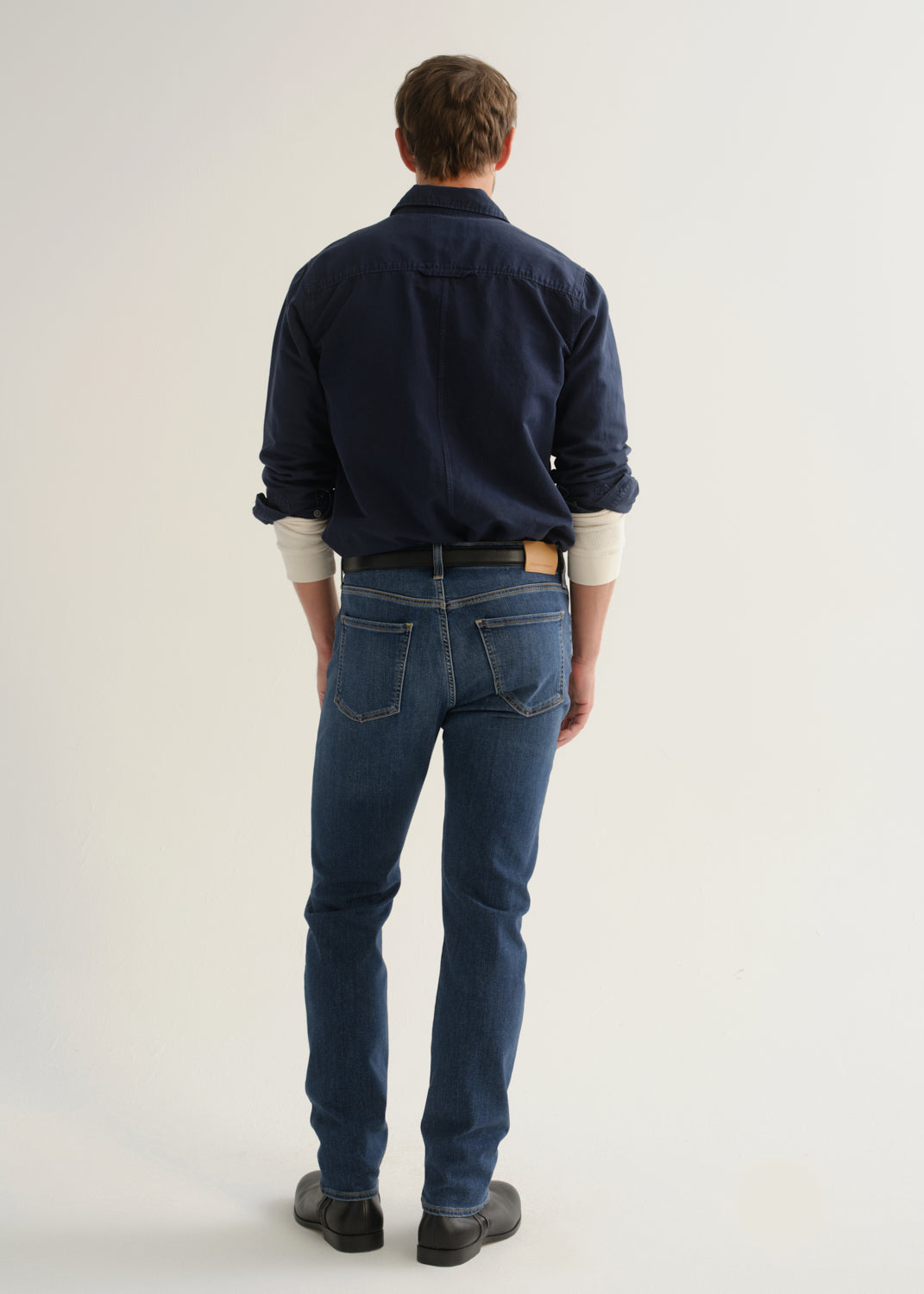 Person wearing medium blue straight leg jeans with a navy long sleeve button-up shirt and black shoes, back full body view on a light grey background.