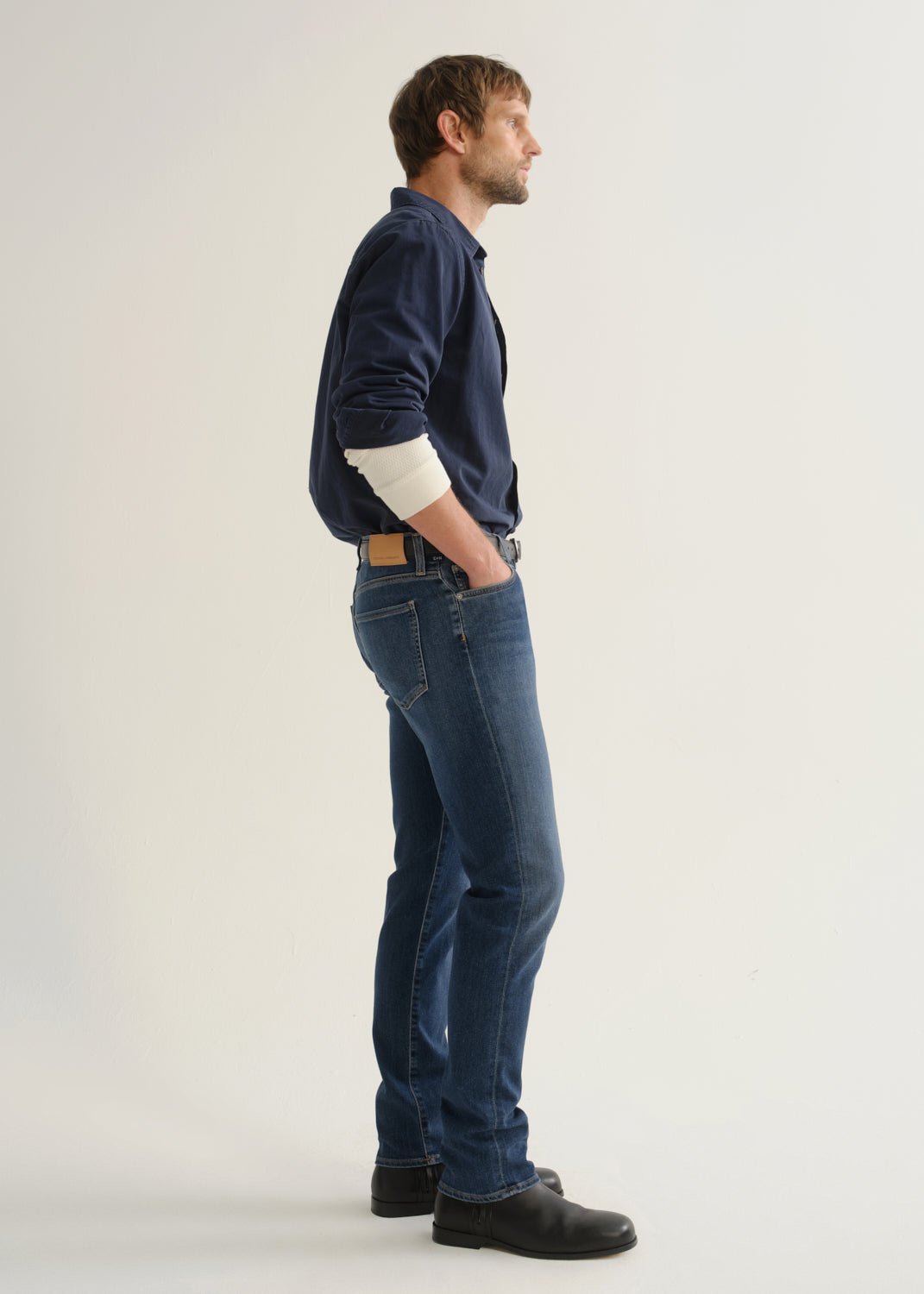 Person wearing medium blue straight leg jeans with a navy long sleeve button-up shirt and black shoes, side full body view on a light grey background.