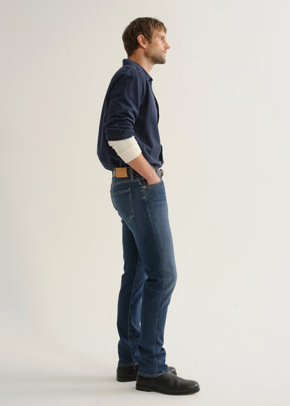 Person wearing medium blue straight leg jeans with a navy long sleeve button-up shirt and black shoes, side full body view on a light grey background.