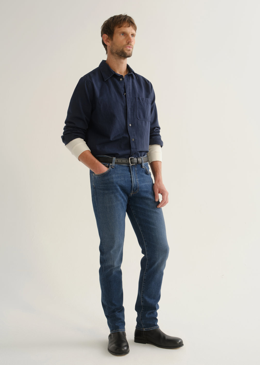 Person wearing medium blue straight leg jeans with a navy long sleeve button-up shirt and black shoes, angled front full body view on a light grey background.