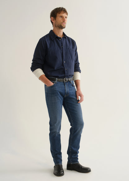 Person wearing medium blue straight leg jeans with a navy long sleeve button-up shirt and black shoes, angled front full body view on a light grey background.